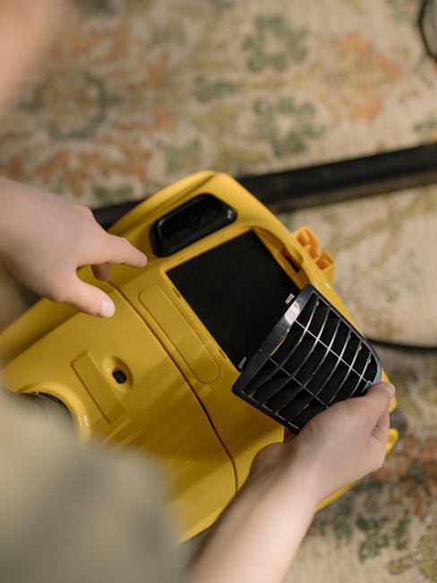 A yellow vacuum cleaner with a black hose and attachment is positioned on a patterned area rug in a living room with wooden flooring. To the left, a black metal and wood media console holds a remote c