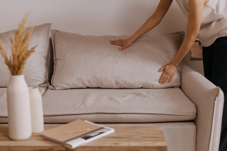 A person dressed in casual clothing is smoothing a beige cushion on a light-colored fabric sofa in a living room. The sofa, which appears to be in a domestic setting, features soft, textured upholster