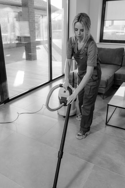 A woman wearing casual clothing and an apron is using a vacuum cleaner with a long hose attachment to clean a tiled floor in a modern, well-lit living room. The room features large glass sliding doors